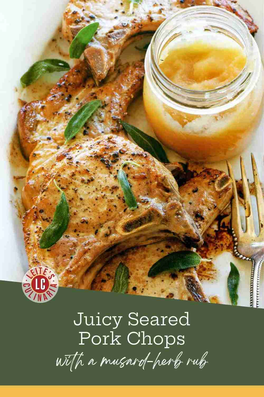 Golden pan-seared pork chops arranged on a white serving platter, garnished with fresh sage leaves and served next to a jar of chunky applesauce.