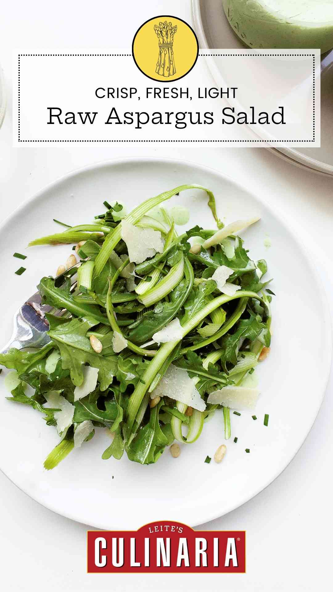 A plate of raw asparagus salad with a jar of dressing and a bowl of pine nuts, and a glass of white wine on the side.