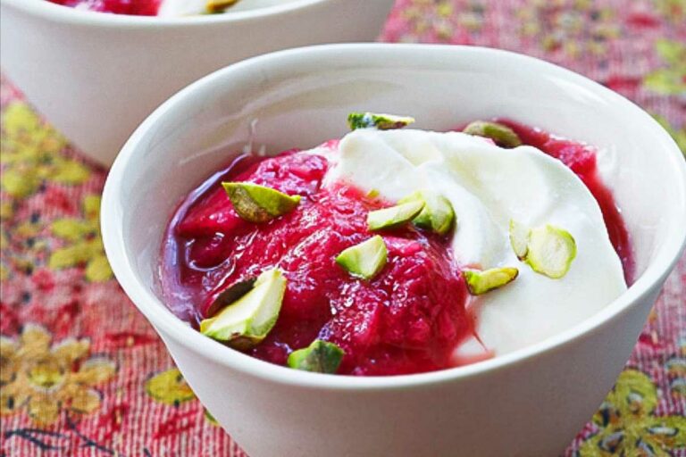 A white bowl of rhubarb and pistachios over thick yogurt on a patterned tablecloth.