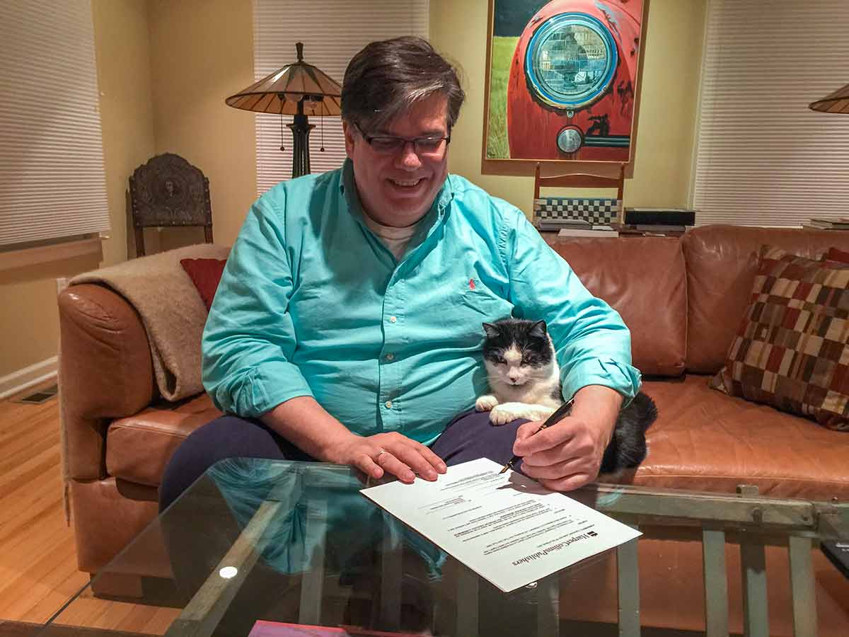 David in an aqua button down shirt with Rory the cat under his left arm, while David is signing the contract for his memoir on a leather couch.