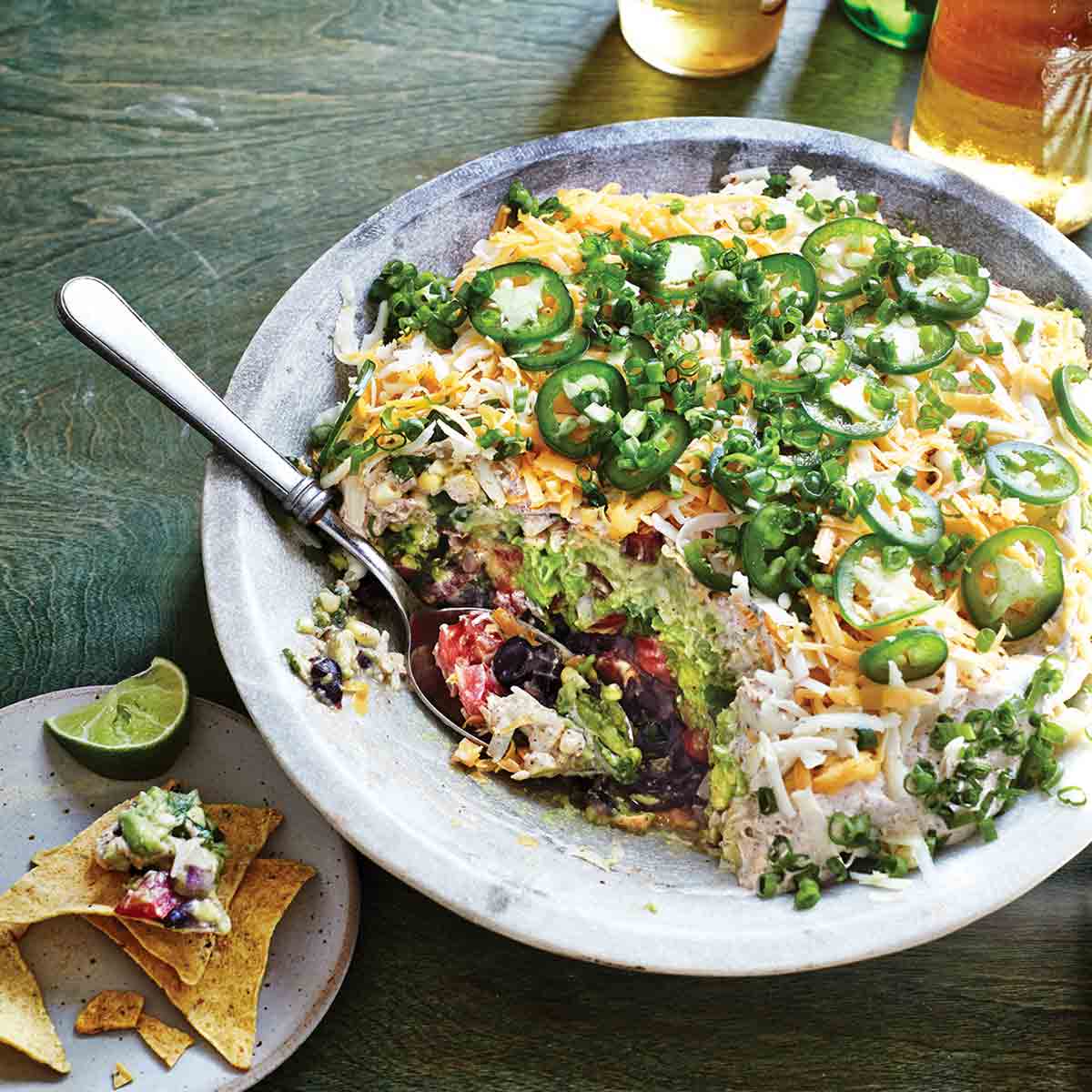 A bowl filled with seven-layer dip with a spoon resting inside and a plate with tortilla chips, dip, and a lime wedge on the side.