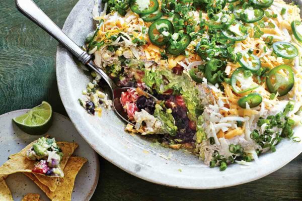 A bowl filled with seven-layer dip with a spoon resting inside and a plate with tortilla chips, dip, and a lime wedge on the side.