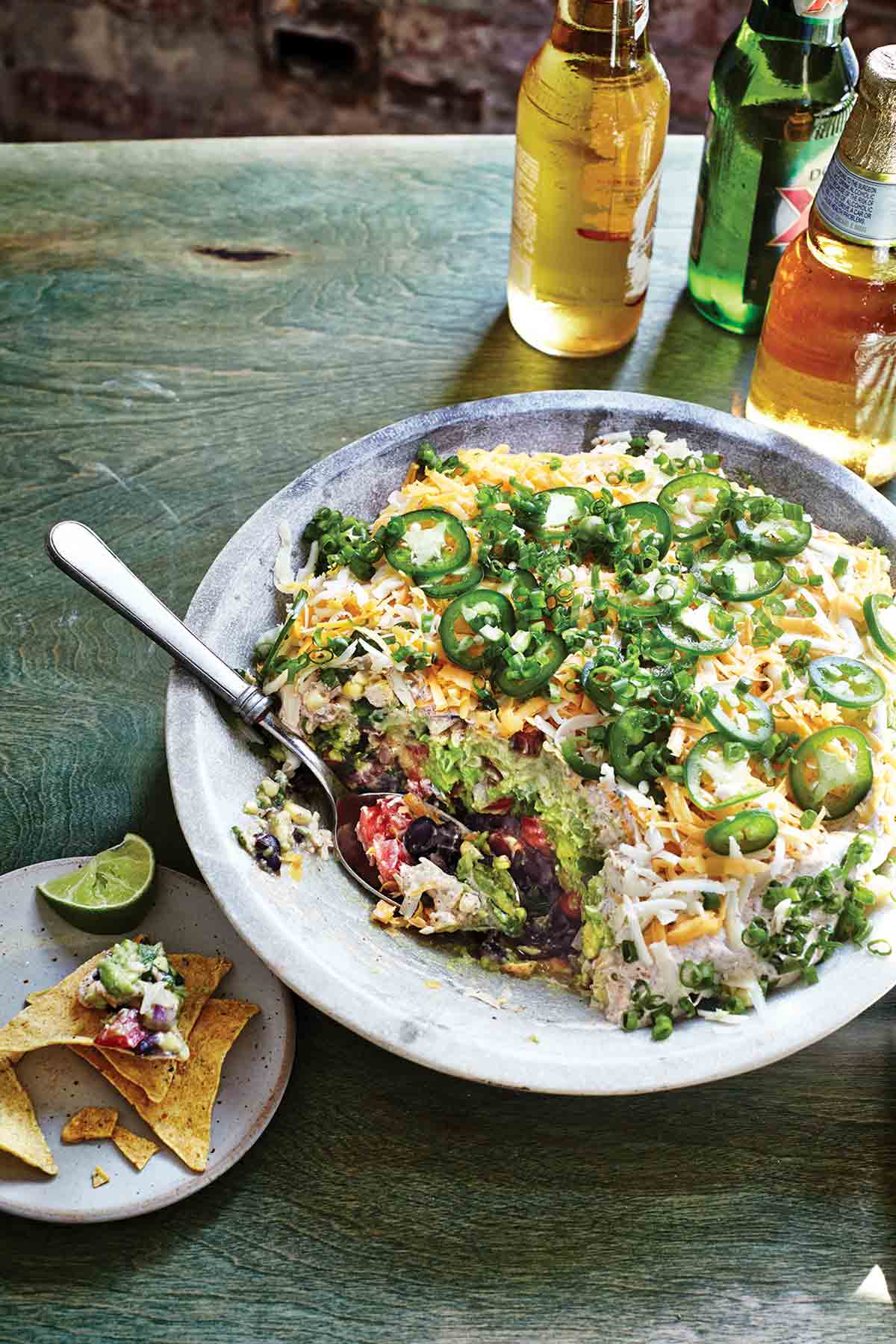 A bowl filled with seven-layer dip with a spoon resting inside and a plate with tortilla chips, dip, and a lime wedge on the side.