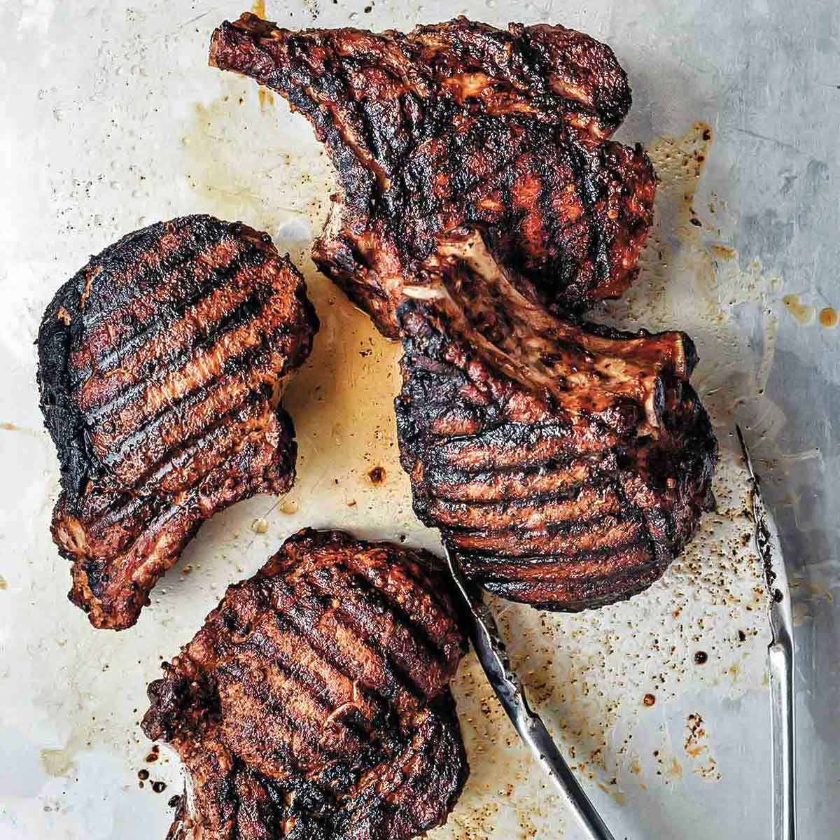 Four spice-rubbed grilled pork chops on a baking sheet with a pair of metal tongs