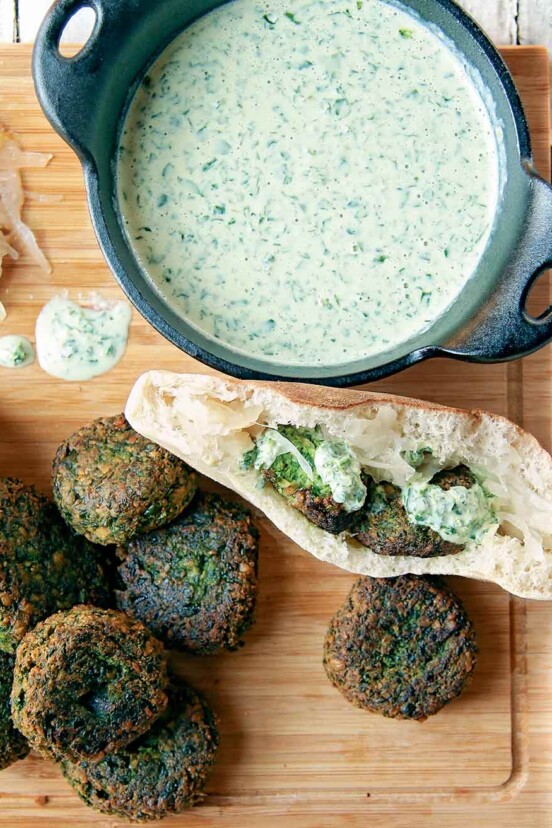 Spinach falafel on a cutting board with some tucked into a pita, and a bowl of parsley tahini sauce.