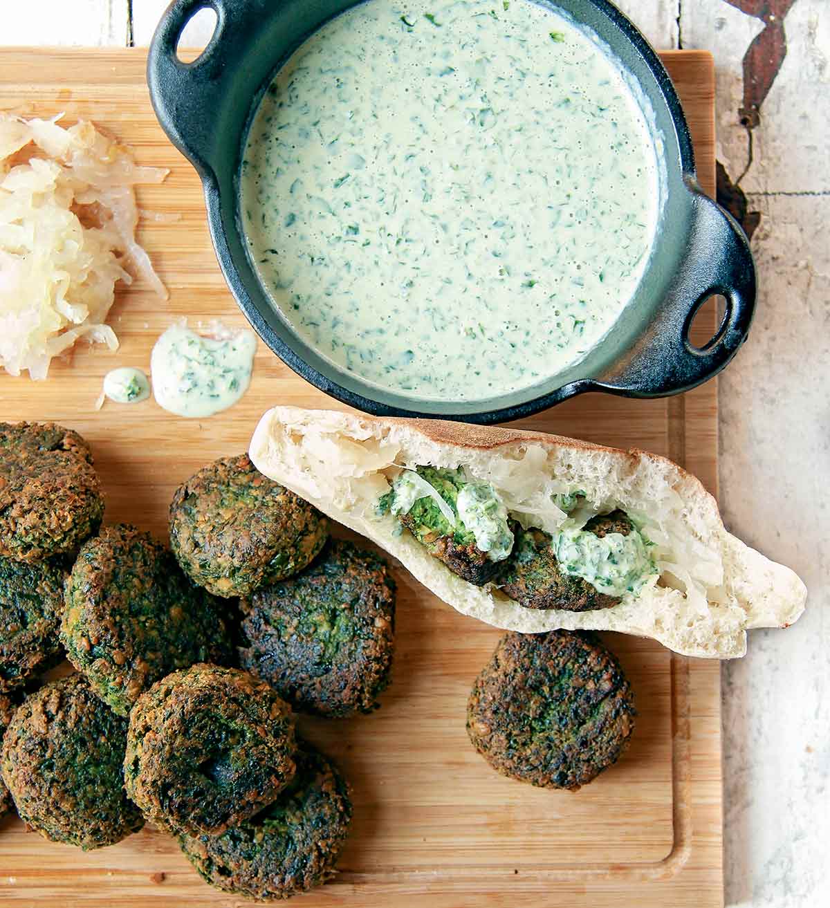 Spinach falafel on a cutting board with some tucked into a pita, and a bowl of parsley tahini sauce.