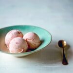A blue bowl with three scoops of strawberry buttermilk ice cream and a gold spoon on the side.