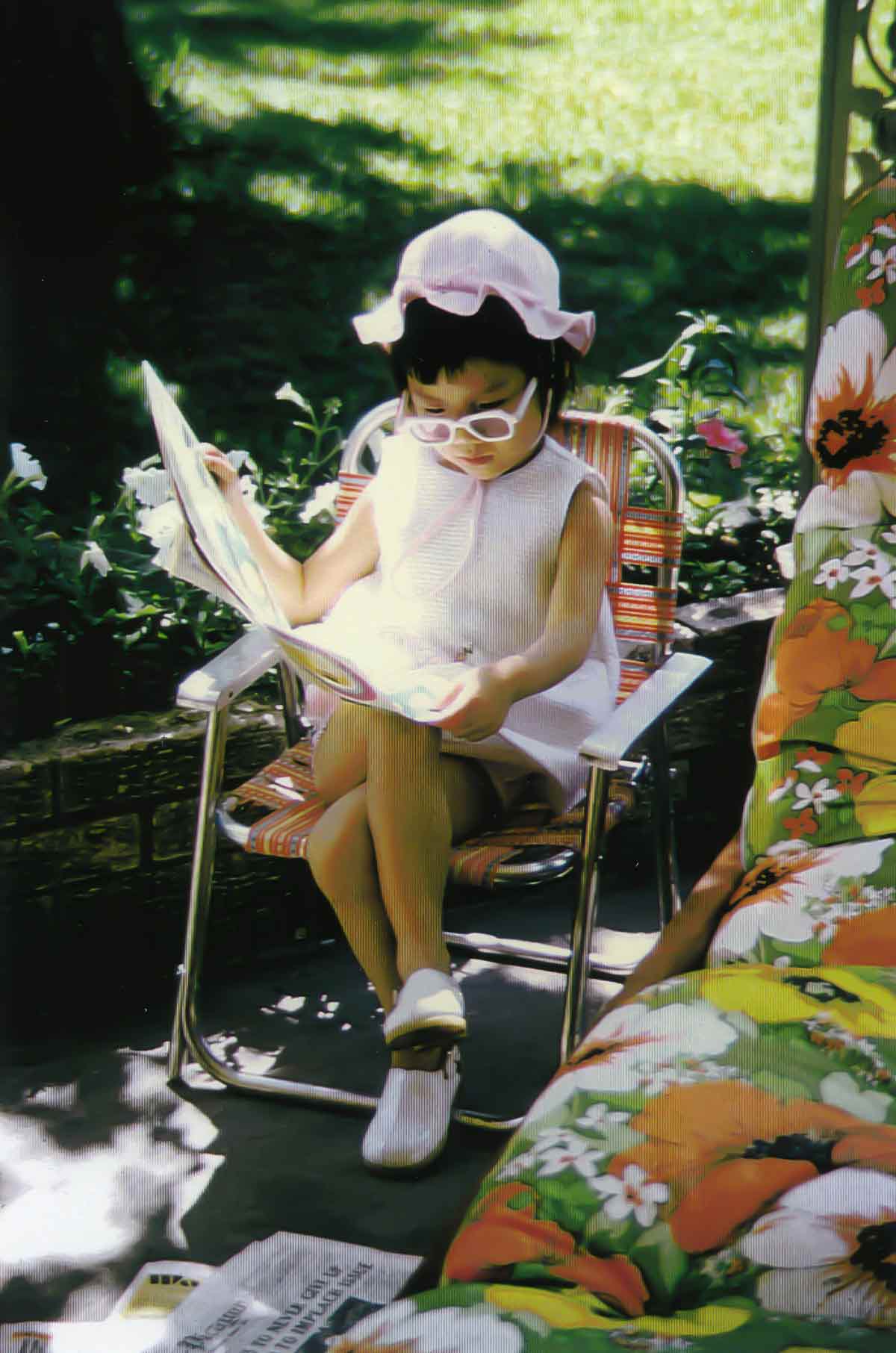 Little girl wearing a hat and sunglasses with legs crossed, sitting outside on a patio chair.