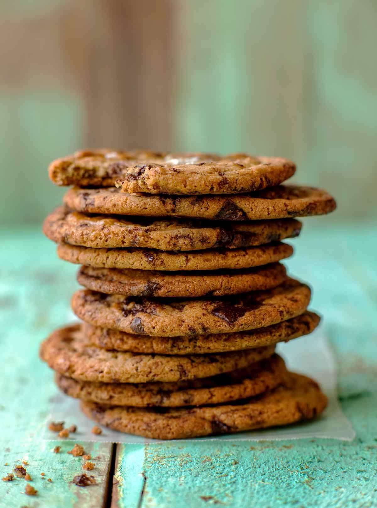 Stack of 10 chocolate chip cookies, each topped with sea salt, on a teal piece of wood