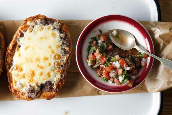Two people reaching for traditional molletes on a platter with a bowl of salsa in the middle.