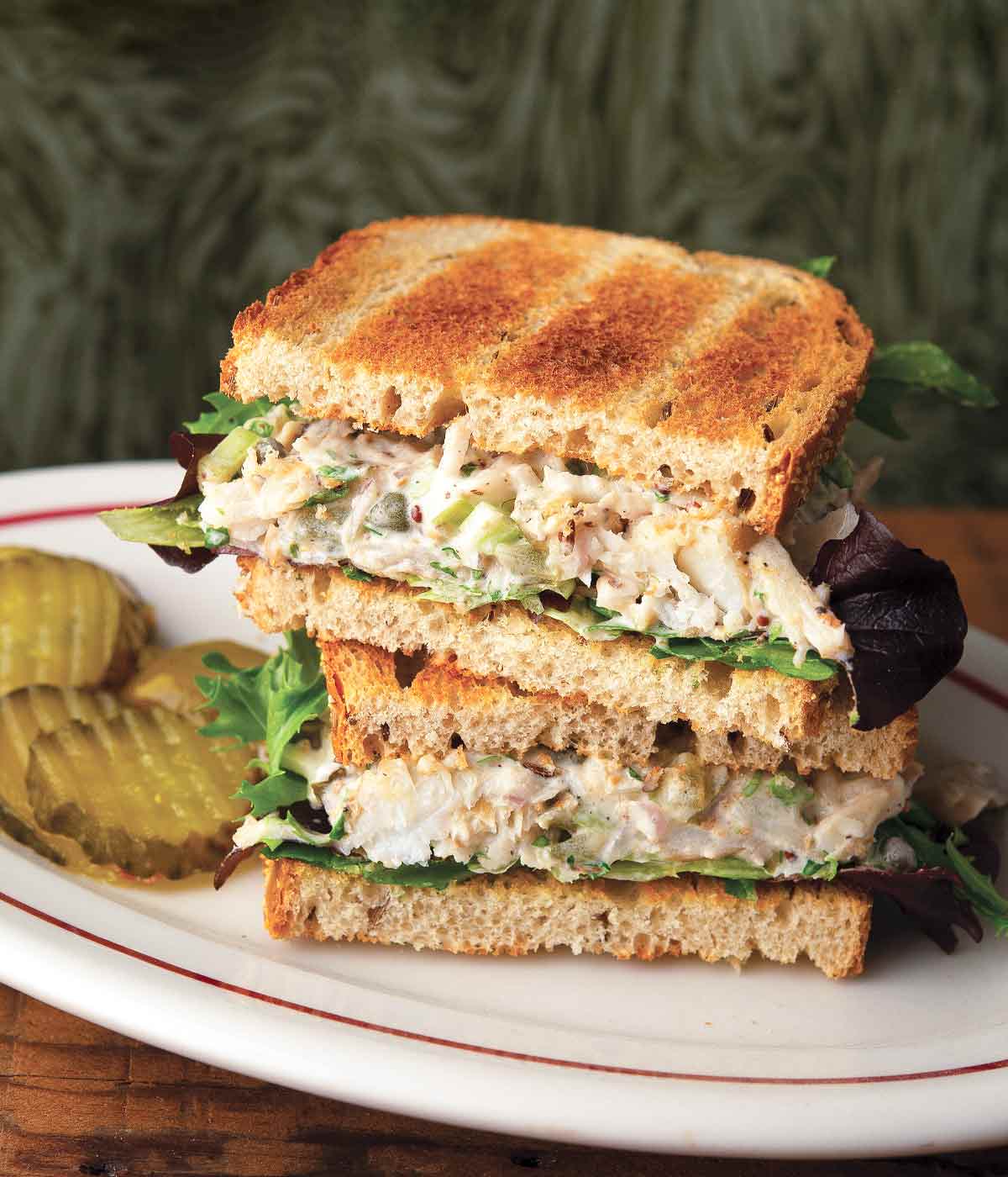 Two halves of a tuna salad sandwich stacked on top of each other with some sliced pickles on the side on a white oval plate.