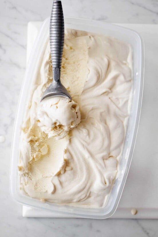 A plastic container filled with vanilla bean ice cream with a metal ice cream scoop resting in it.