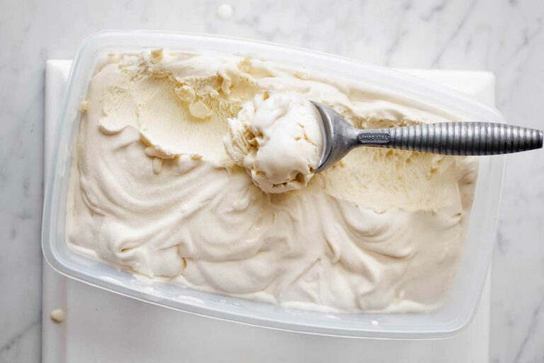 A plastic container filled with vanilla bean ice cream with a metal ice cream scoop resting in it.