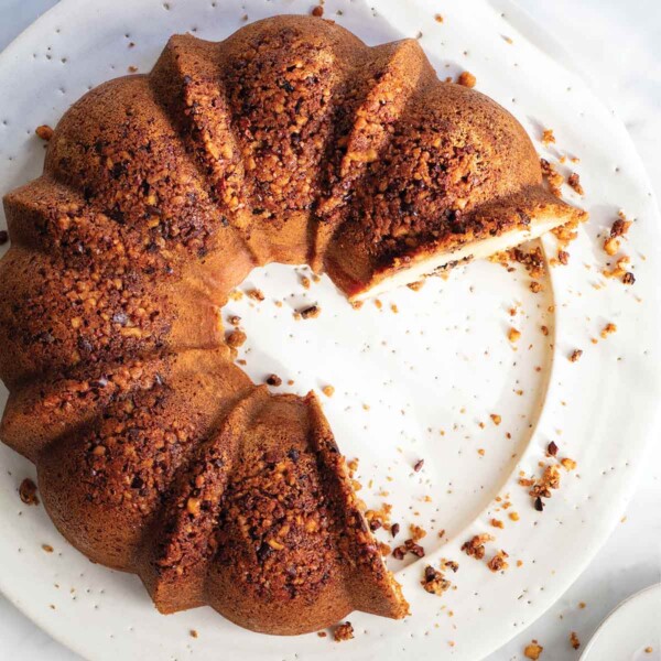 A Bundt-shaped gluten-free walnut coffee cake with cacao nibs on a white platter with several slices missing.