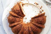 A Bundt-shaped gluten-free walnut coffee cake with cacao nibs on a white platter with several slices missing.