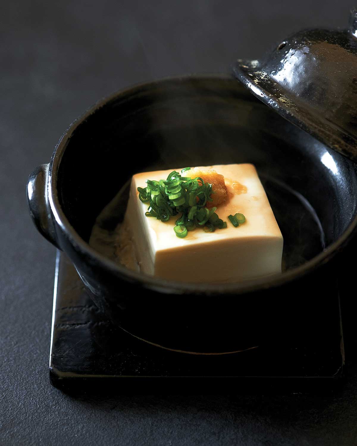 A black bowl with a block of warm tofu with soy sauce, ginger, and scallion garnish.
