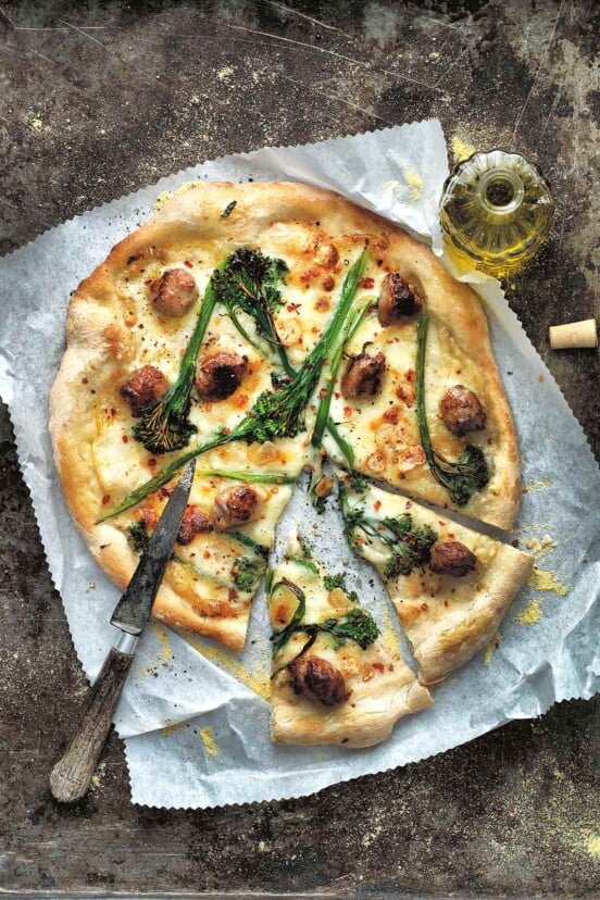 A white pizza with broccolini, fontina, and sausage cut into slices on a piece of parchment with a knife on top and a bottle of oil on the side.