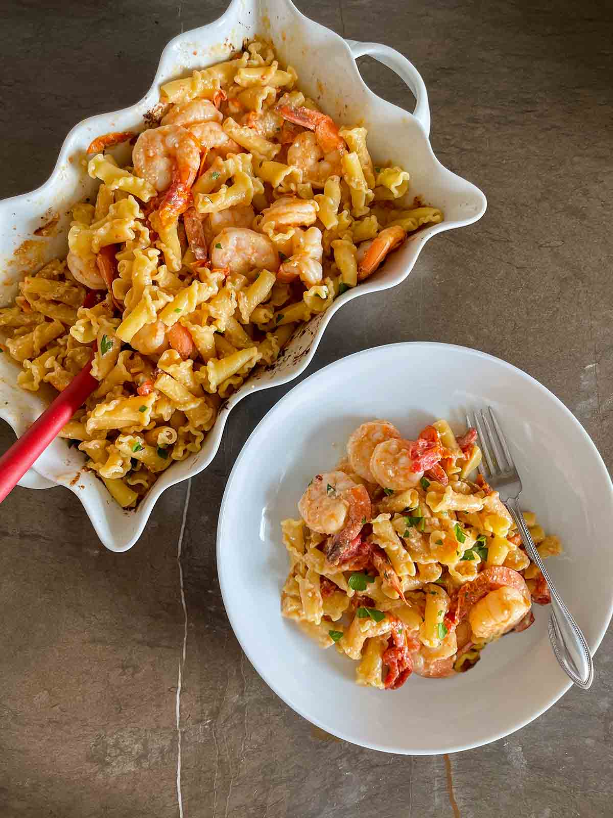 En serveringsfat och skål båda fyllda med bakad pasta med räkor, fetaost och tomater, med en gaffel på sidan.