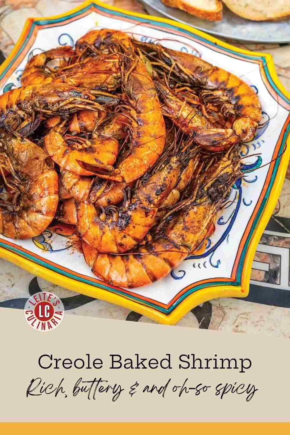 A platter of baked jumbo shrimp coated in a rich, dark Creole sauce next to a plate of sliced crusty bread.