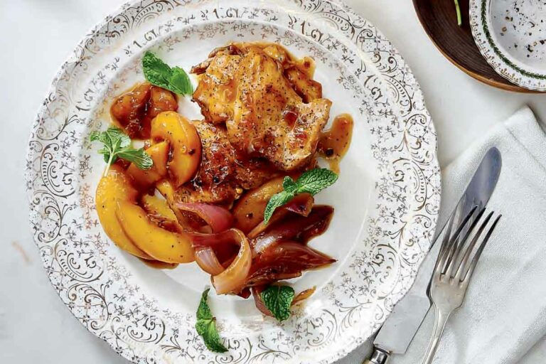 A patterned plate topped with chicken thighs with spicy peach sauce on a table with a bottle and glass of wine, flowers, cutlery, and a napkin.