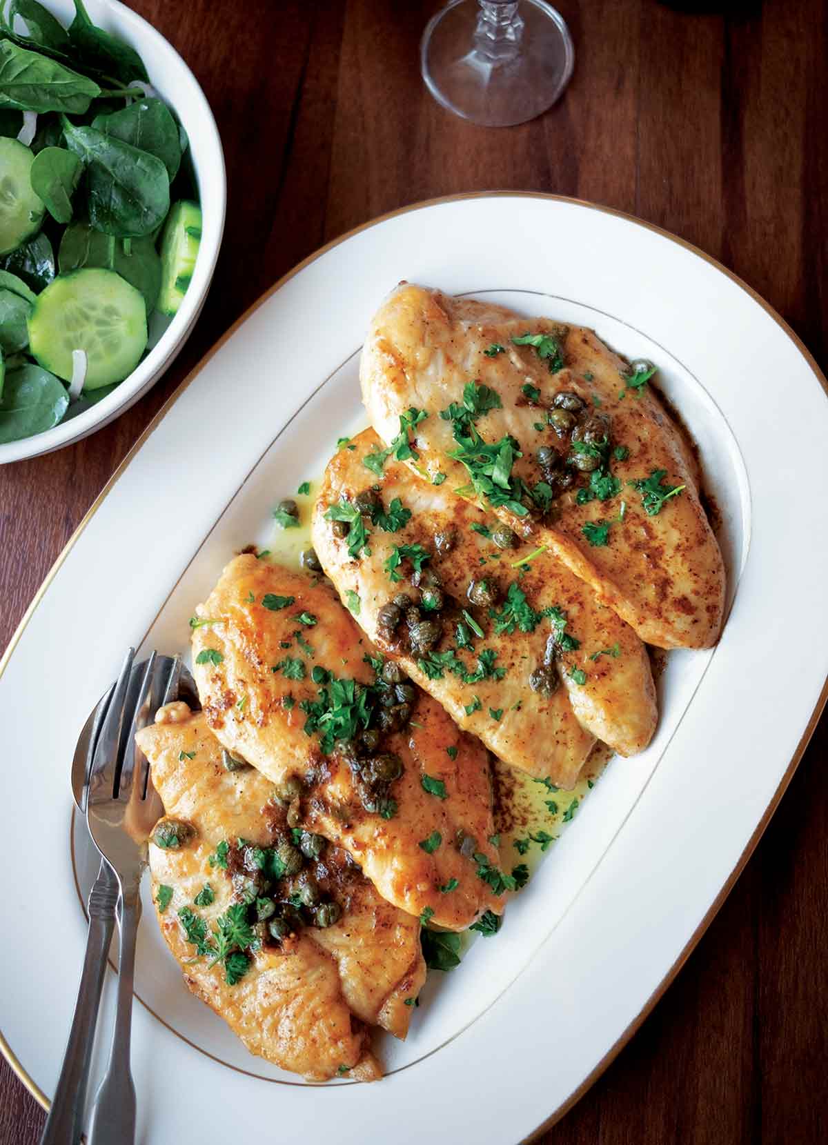 Four pieces of classic chicken piccata topped with parsley and capers on a white oval serving plate.