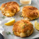 Three crab cakes with ritz crackers on a metal tray with a small bowl of lemon butter, lemon wedges, and dill sprigs nearby.