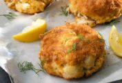Three crab cakes with ritz crackers on a metal tray with a small bowl of lemon butter, lemon wedges, and dill sprigs nearby.