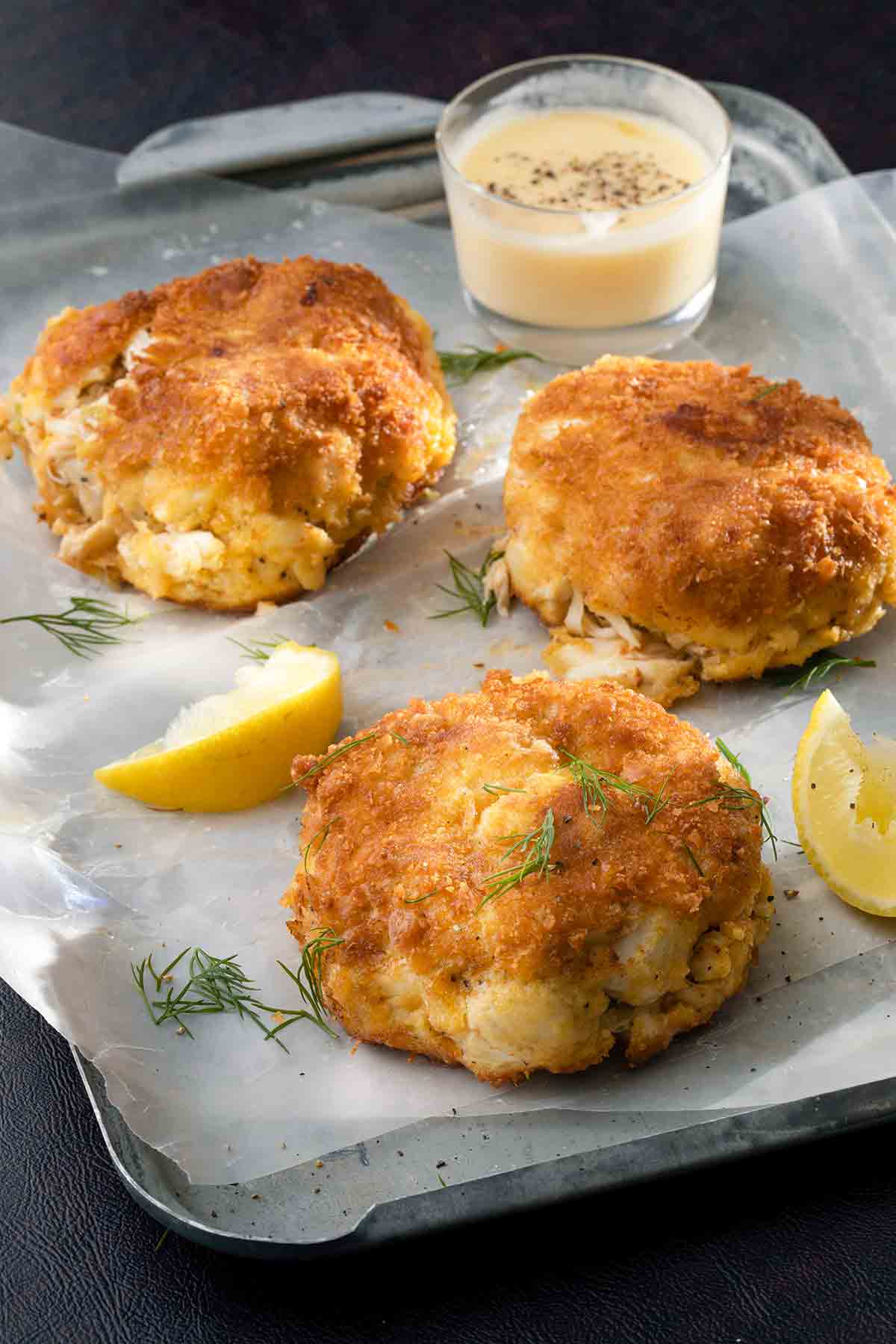 Three crab cakes with ritz crackers on a metal tray with a small bowl of lemon butter, lemon wedges, and dill sprigs nearby.