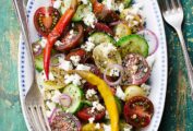 An oval platter topped with Greek salad - sliced cucumber, tomatoes, red onion, peppers, and feta cheese.