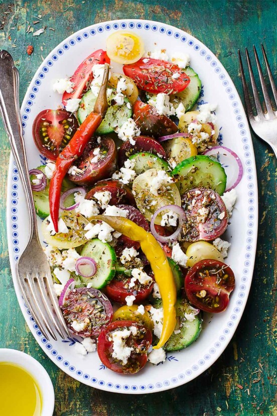 An oval platter topped with Greek salad - sliced cucumber, tomatoes, red onion, peppers, and feta cheese.