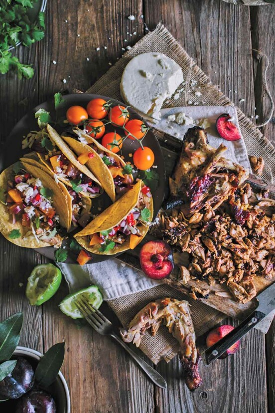 A black platter with several grilled chicken tacos with fruit salsa, a few cherry tomatoes, and a wooden board with cut chicken, halved plums, and queso fresco.