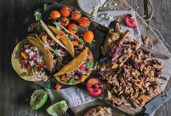 A black platter with several grilled chicken tacos with fruit salsa, a few cherry tomatoes, and a wooden board with cut chicken, halved plums, and queso fresco.