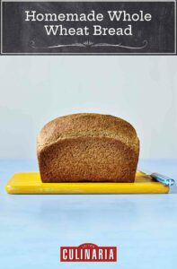 A loaf of unsliced whole wheat bread sitting on a yellow cutting board with a blue-handled knife beside it
