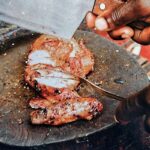 A sliced jerk chicken breast on a round cutting board, with a man's hand holding a cleaver above it