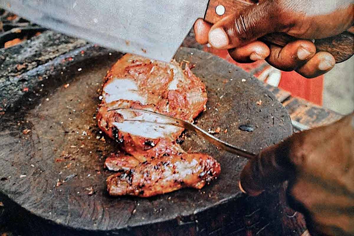 A sliced jerk chicken breast on a round cutting board, with a man's hand holding a cleaver above it