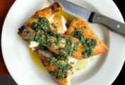A portion of Jonathan Waxman's roast chicken topped with salsa verde on a white plate with a knife on the side.