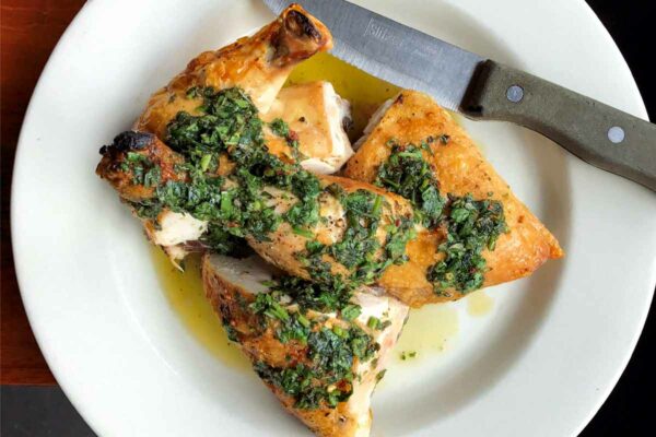 A portion of Jonathan Waxman's roast chicken topped with salsa verde on a white plate with a knife on the side.