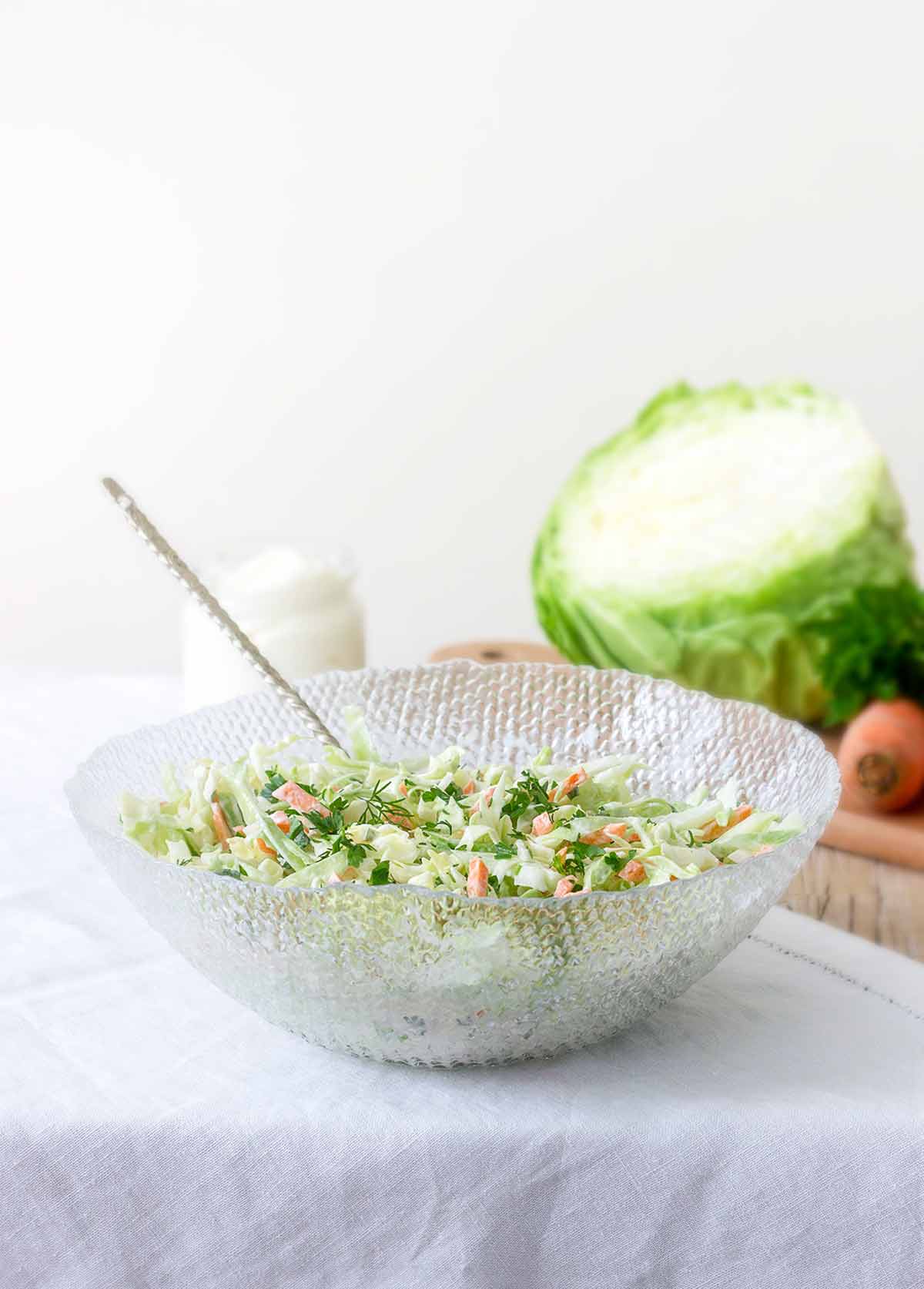 Glass bowl with lime coleslaw--green cabbage, carrots, lime zest, mayo, and parsley--on a tablecloth, cabbage and carrots behind