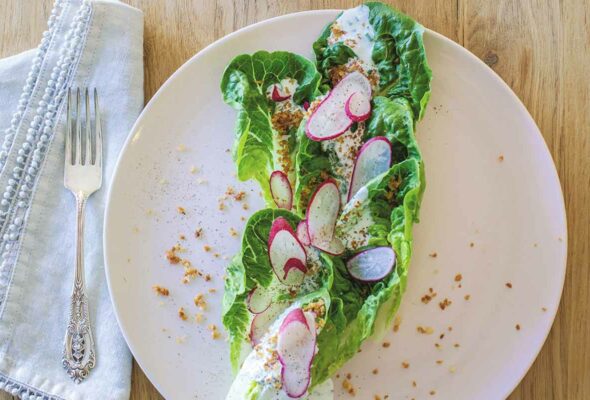 A little gem salad with bread crumb croutons and sliced radishes on a white plate with a bowl of dressing nearby.