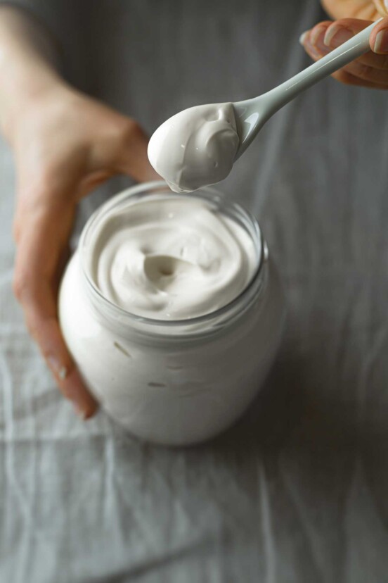 A person holding a spoonful of milk mayonnaise above a jar.