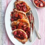 Four pork chops with strawberry balsamic sauce on top arranged on a white oval platter with serving utensils and a bowl of strawberry sauce on the side.