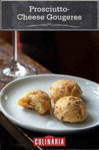 A white plate with three prosciutto-cheese gougeres with a glass of Prosecco in the background.
