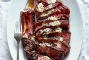 A sliced rib eye on an oval platter with anchovy butter drizzled over the top and a fork resting on the side.