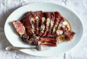 A sliced rib eye on an oval platter with anchovy butter drizzled over the top and a fork resting on the side.