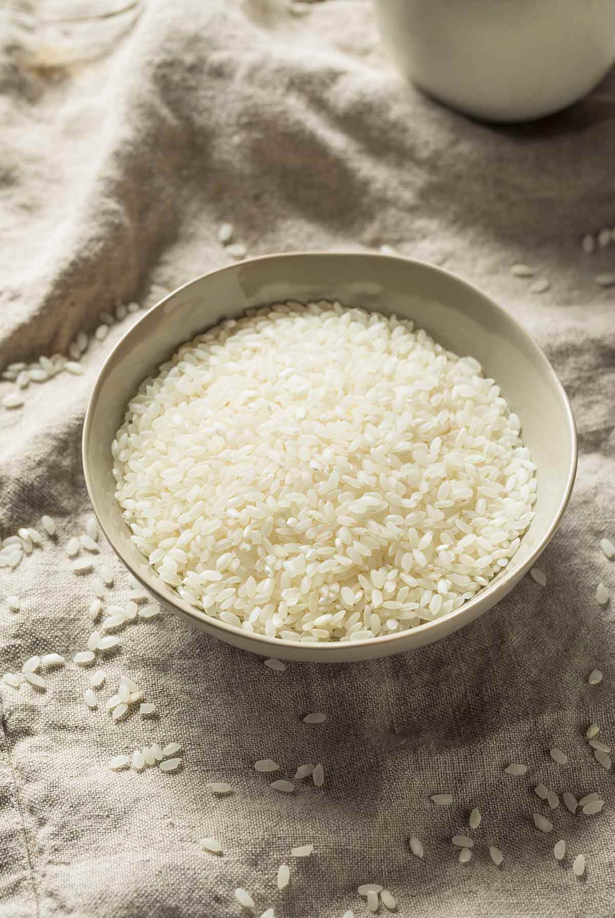 A bowl of white rice with rice grains scattered around it.