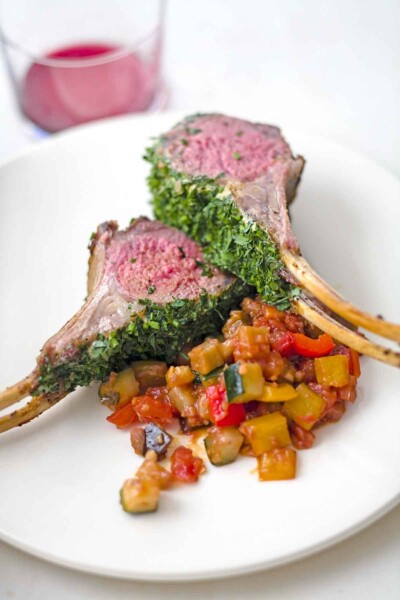 Two chops from a roasted rack of lamb with parsley, Dijon, and chives on a white plate with a cooked zucchini and tomato mixture.