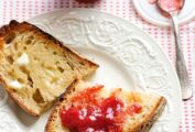 Two pieces of buttered toast, one with strawberry jam, on a white plate with a spoon and jar of jam in the background.