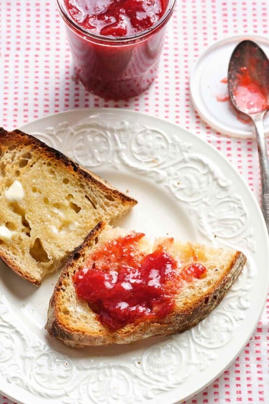 Two pieces of buttered toast, one with strawberry jam, on a white plate with a spoon and jar of jam in the background.