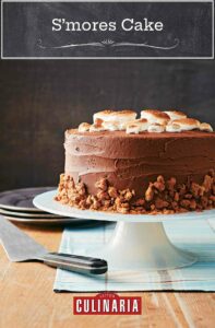 A s'mores cake, topped with toasted marshmallows and graham cracker crumbles on a cake stand.