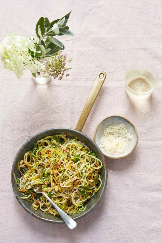 A skillet filled with spaghetti with zucchini, lemon, and basil, with a fork resting in it and a bowl of Parmesan on the side.